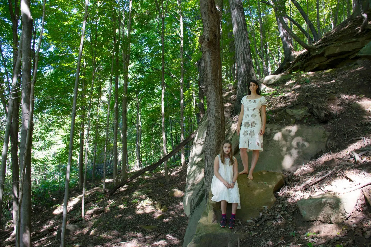 Standing barefoot on a sandstone ledge in a pale dress marked with rust-colored prints, my daughter seated on the rock below me