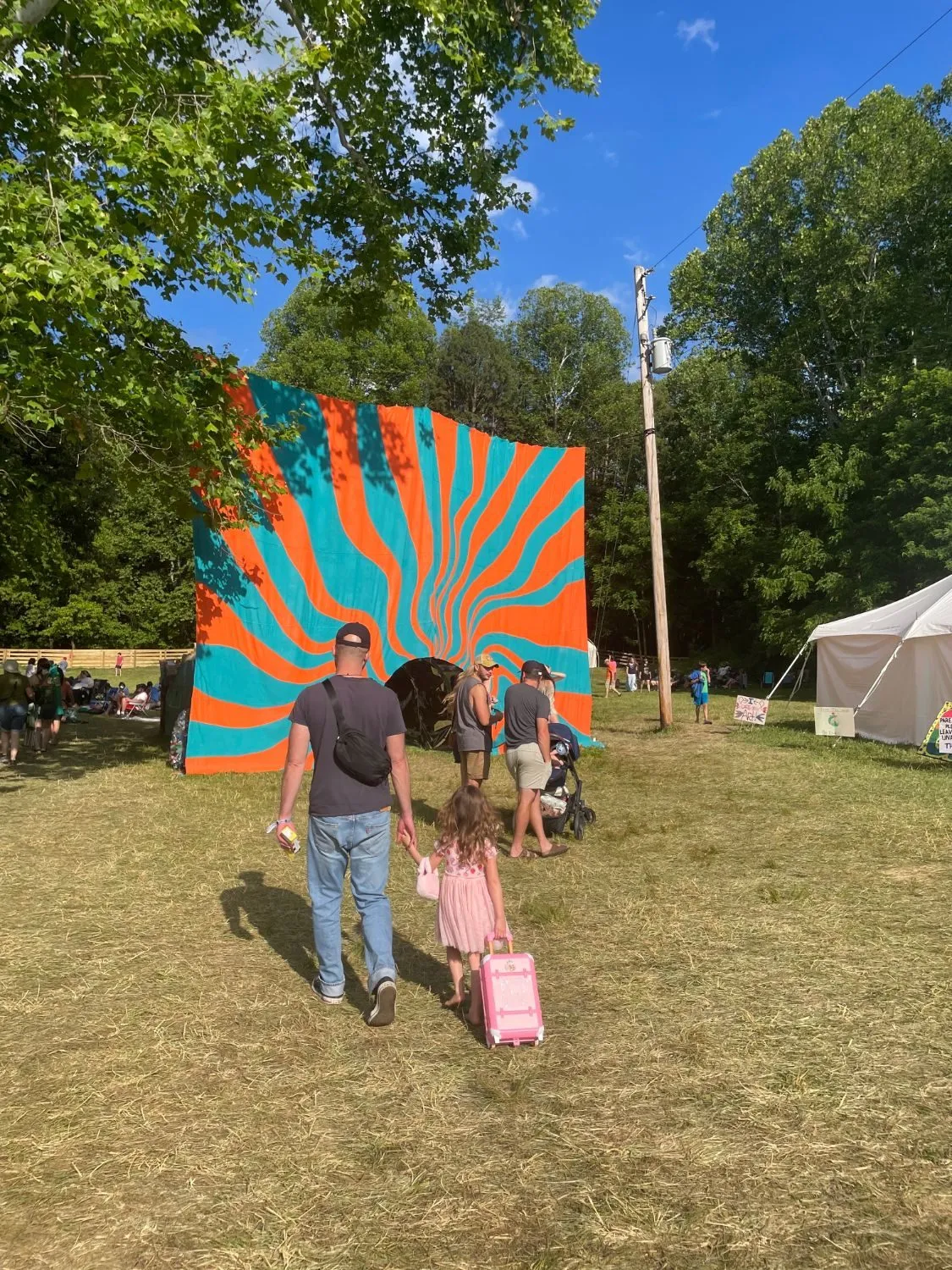 A man and a small girl with a pink suitcase crossing the grass toward the installation under a blue sky