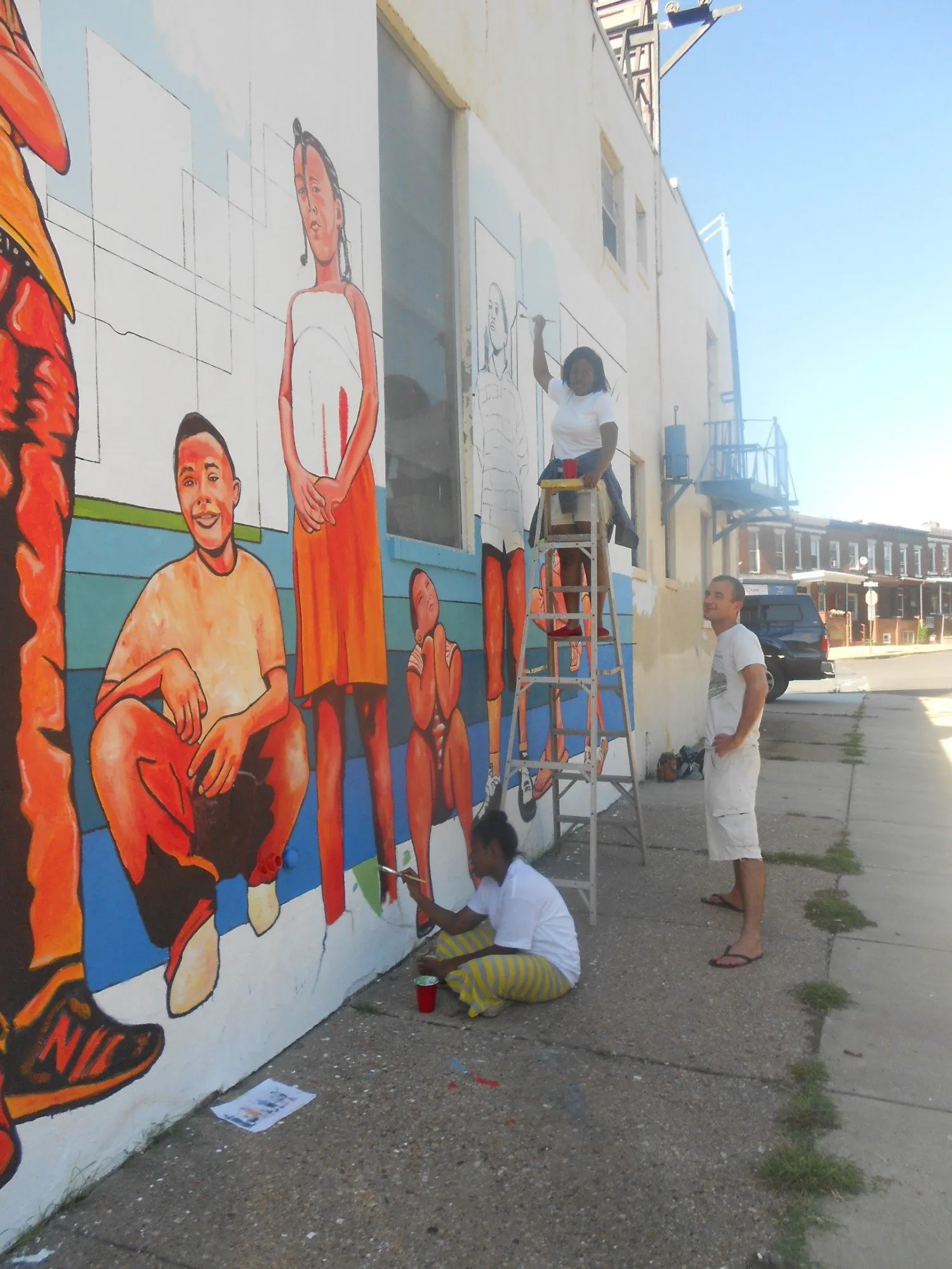 The mural underway on the wall, painted figures finished at the left while a helper on a ladder works on one still in outline