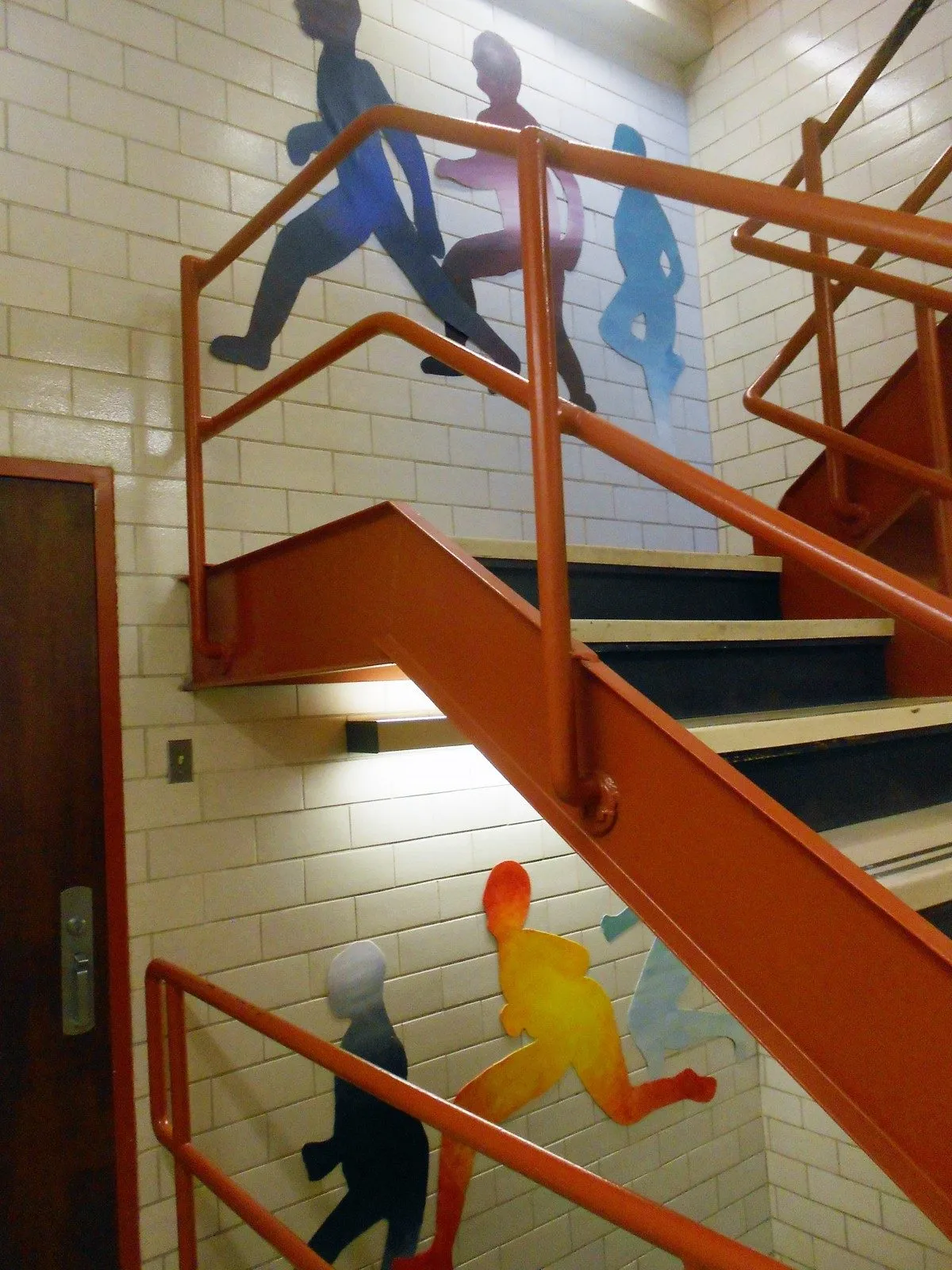 The stairwell seen from a landing, cut metal figures striding across the white tiled wall above and below the orange handrails
