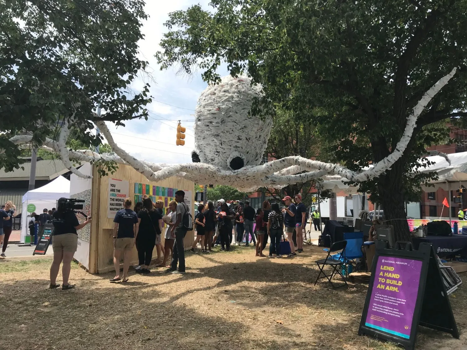 A crowd gathered beneath the octopus in daylight, beside a National Aquarium sign reading "Lend a hand to build an arm"