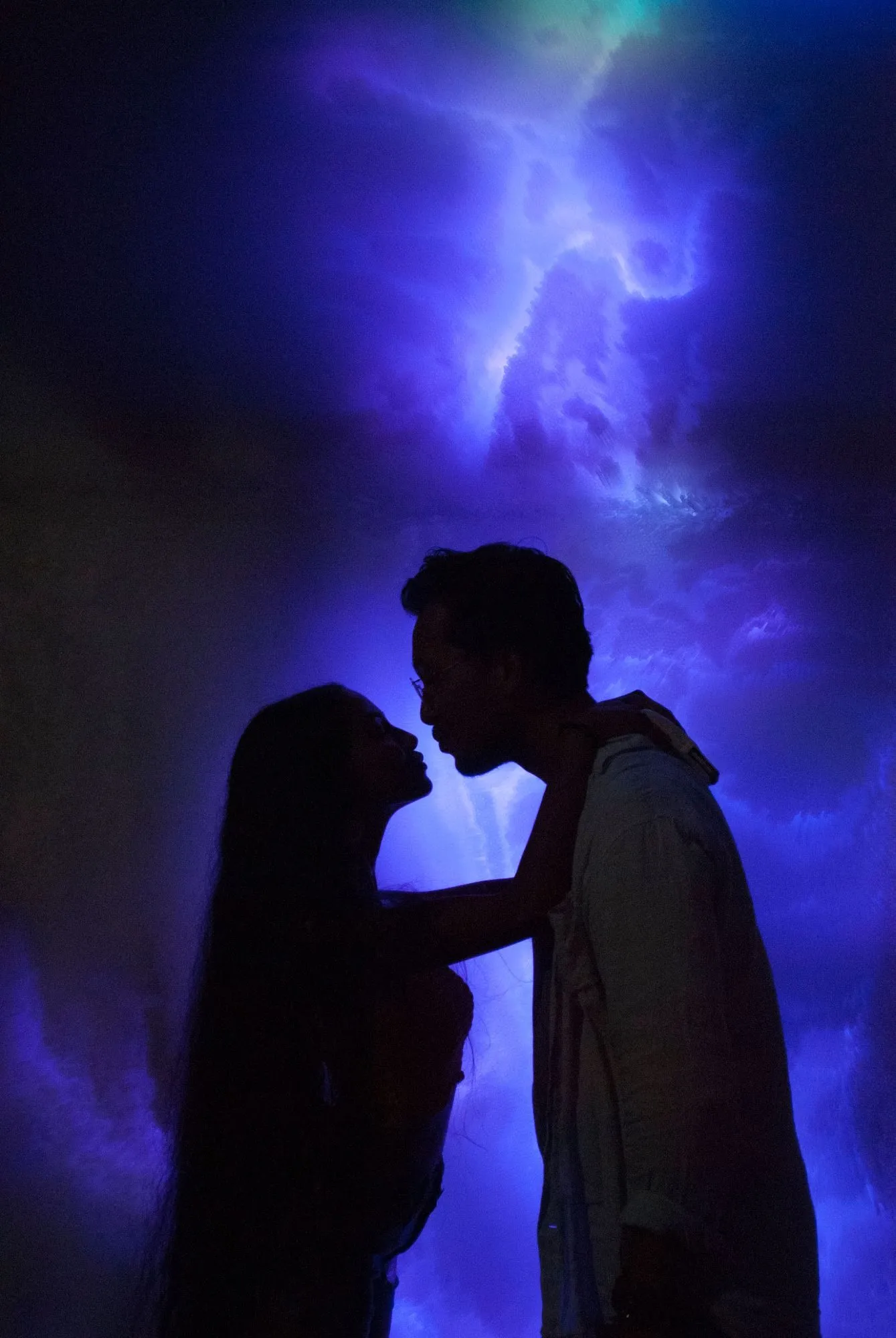 A couple silhouetted face to face against the blue light of the piece