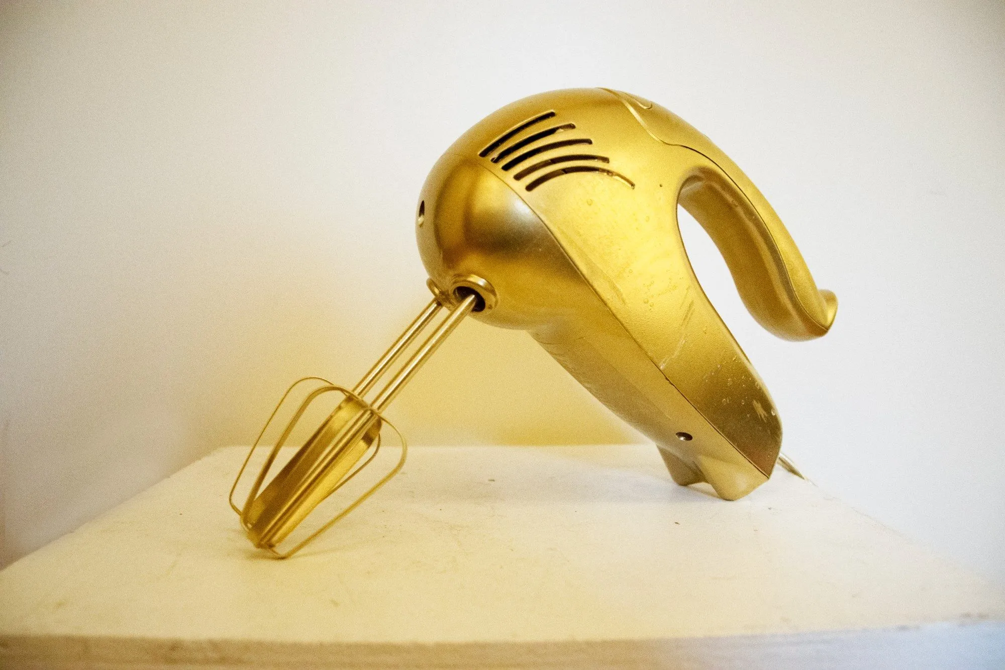 A hand mixer painted gold, beaters attached, resting on a white plinth
