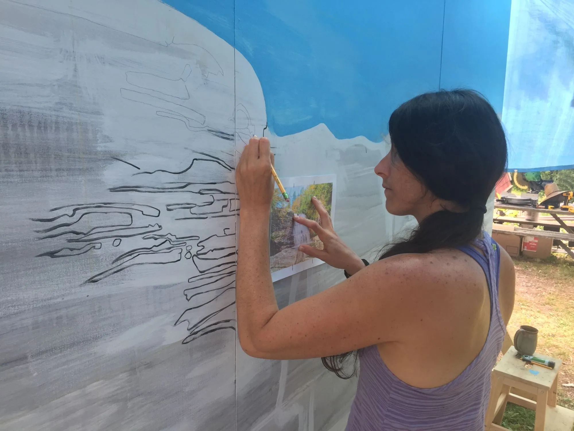 Kasey drawing the rock ledges in pencil over the grey underpainting, a reference print held flat against the wall