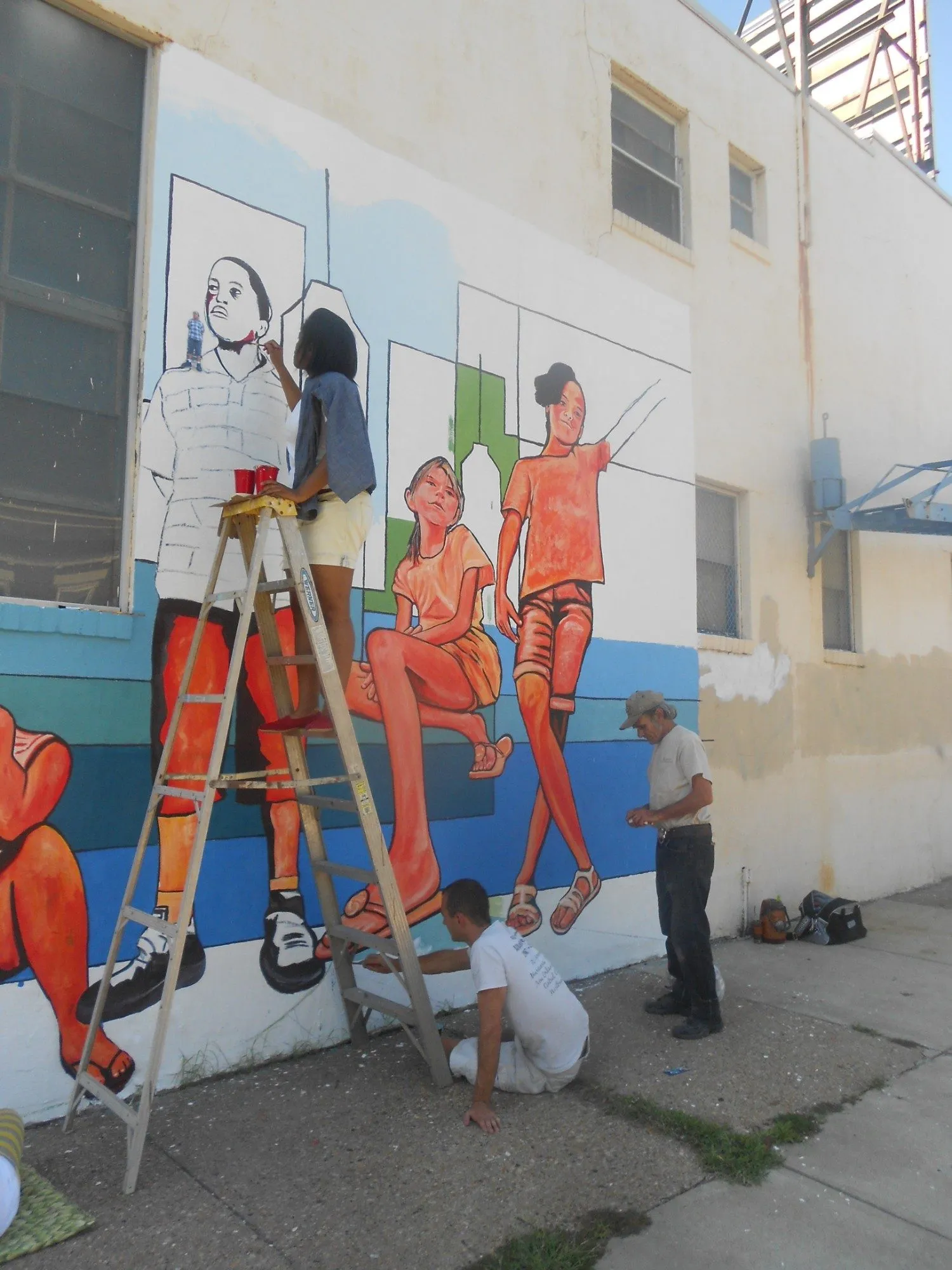 Two orange figures completed against the blue bands of the skyline, with a helper on a ladder painting the outlined figure beside them