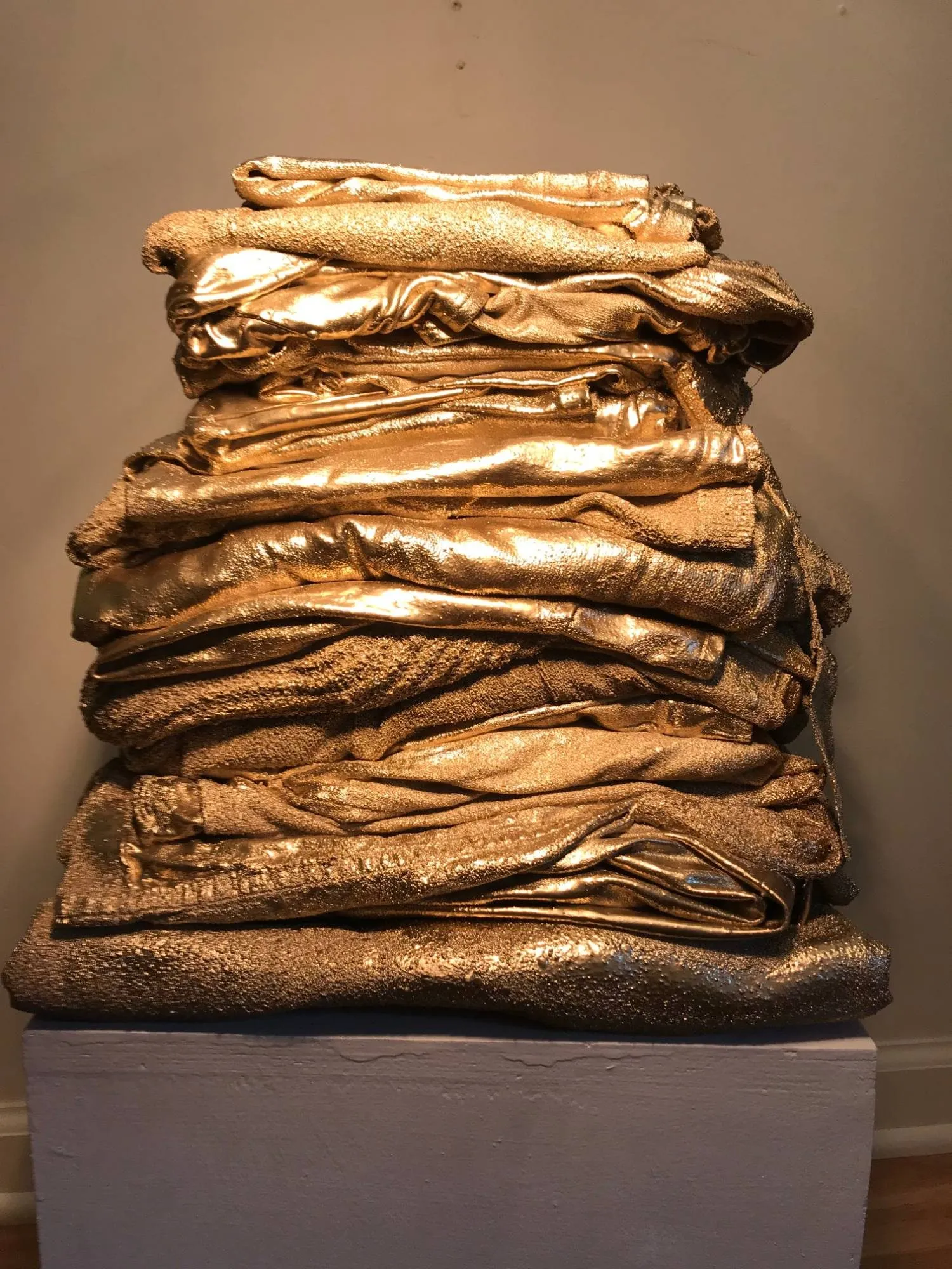 A tall stack of folded laundry, every piece painted gold, sitting on a white plinth