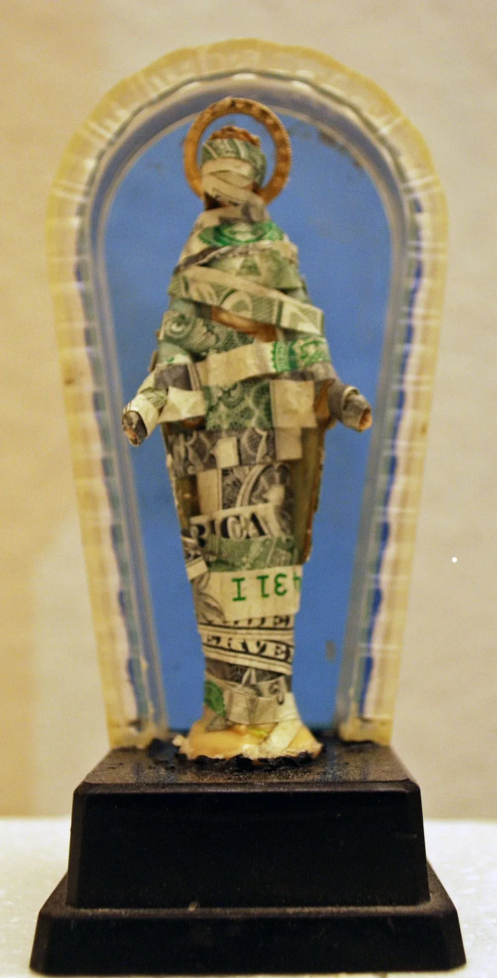 A small standing figure wrapped in dollar bills, a coin halo behind her head, set in a shrine on a black base