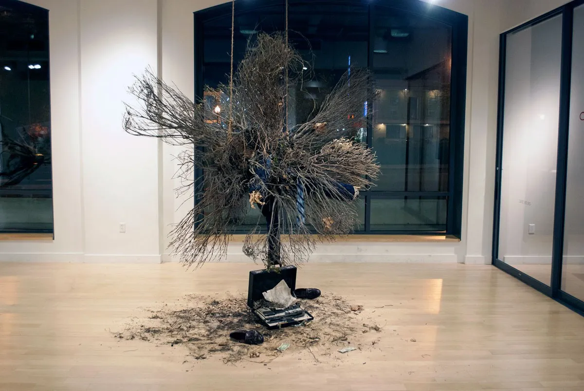 A wide burst of bare branches rising from an open suitcase, debris scattered in a ring across the gallery floor