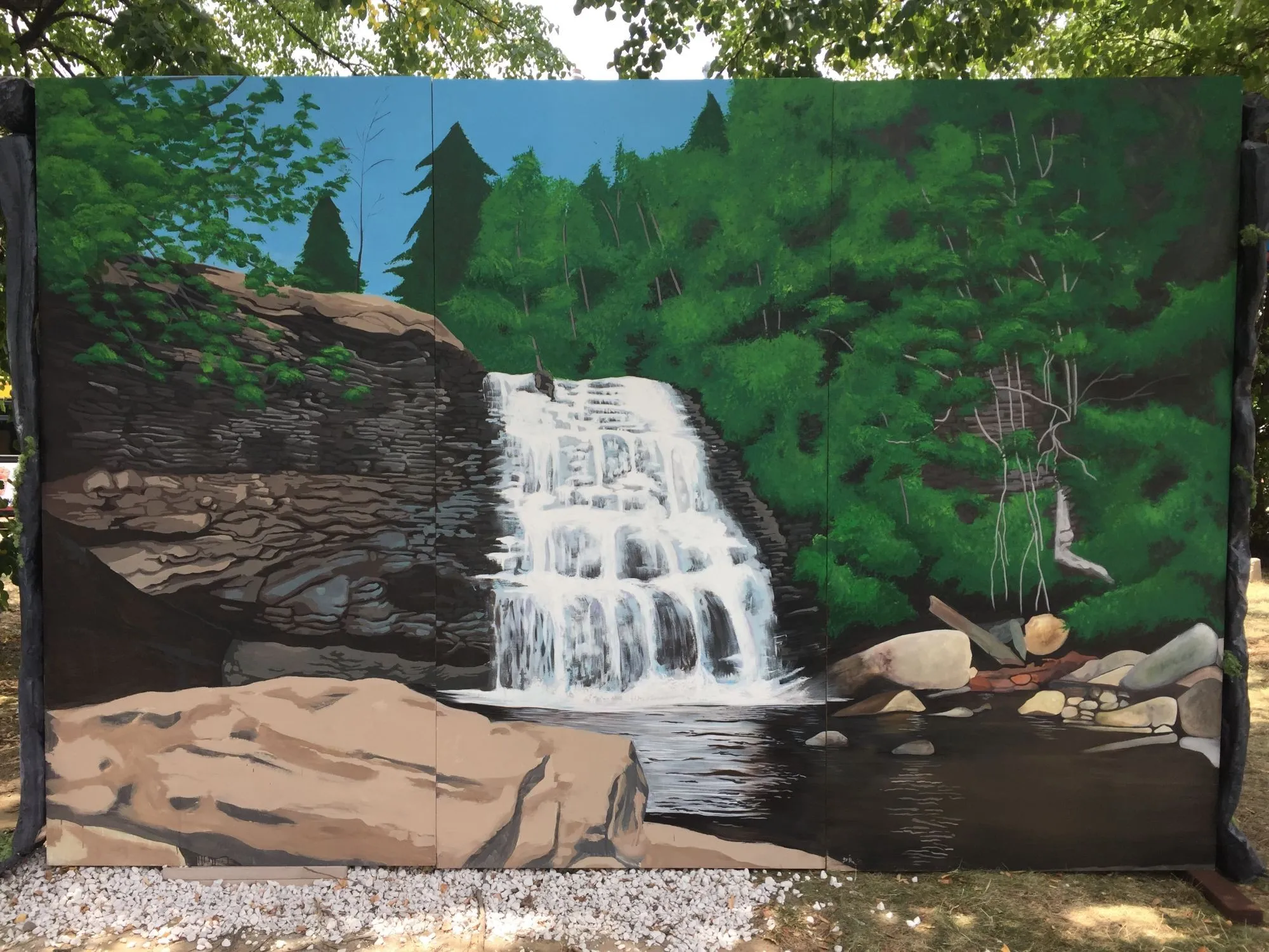 The finished mural standing outdoors on its panels, black-painted posts either side and real trees overhanging it