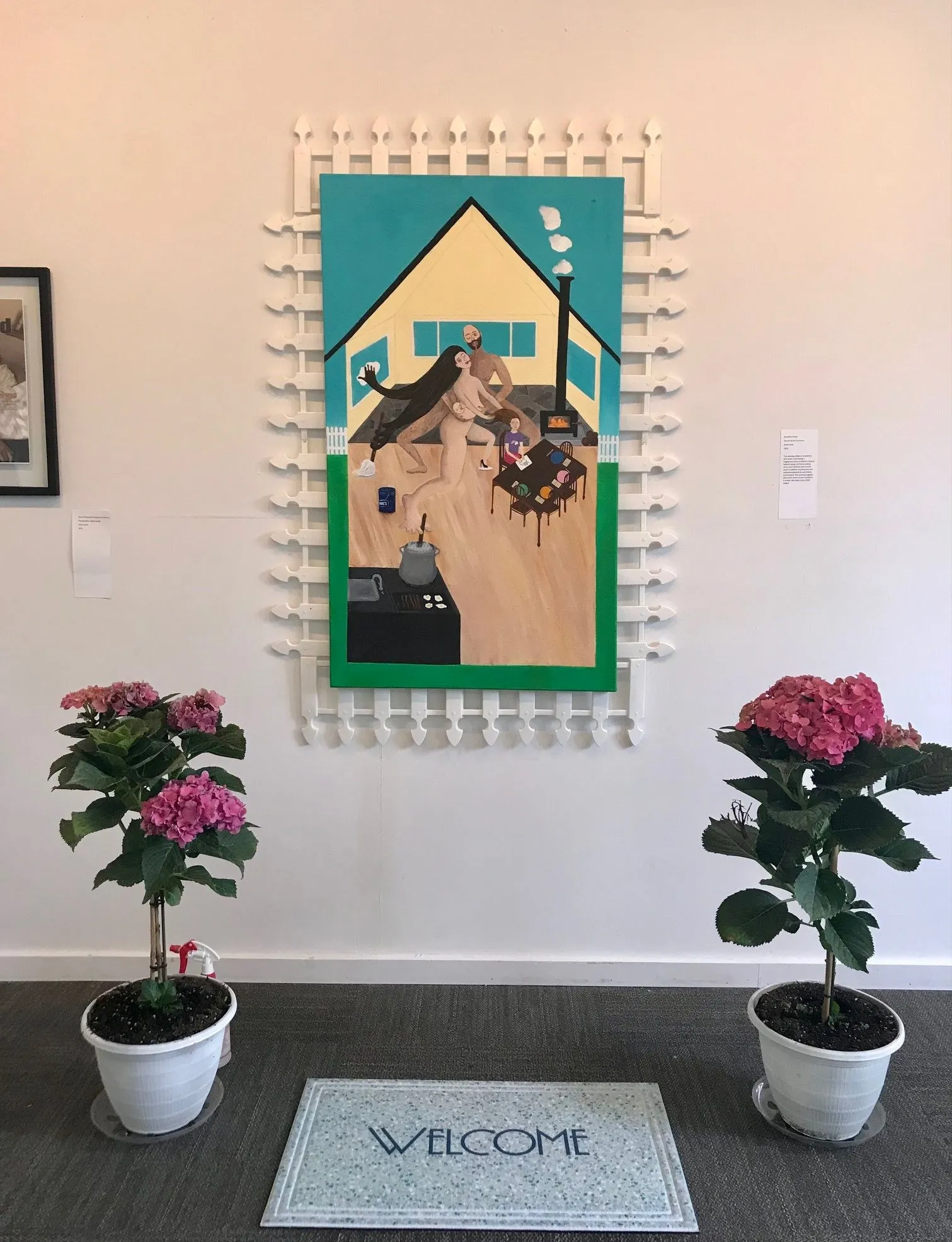 The painting hung on a gallery wall inside a white picket fence frame, its wall label to the right, potted hydrangeas and a welcome mat on the floor below