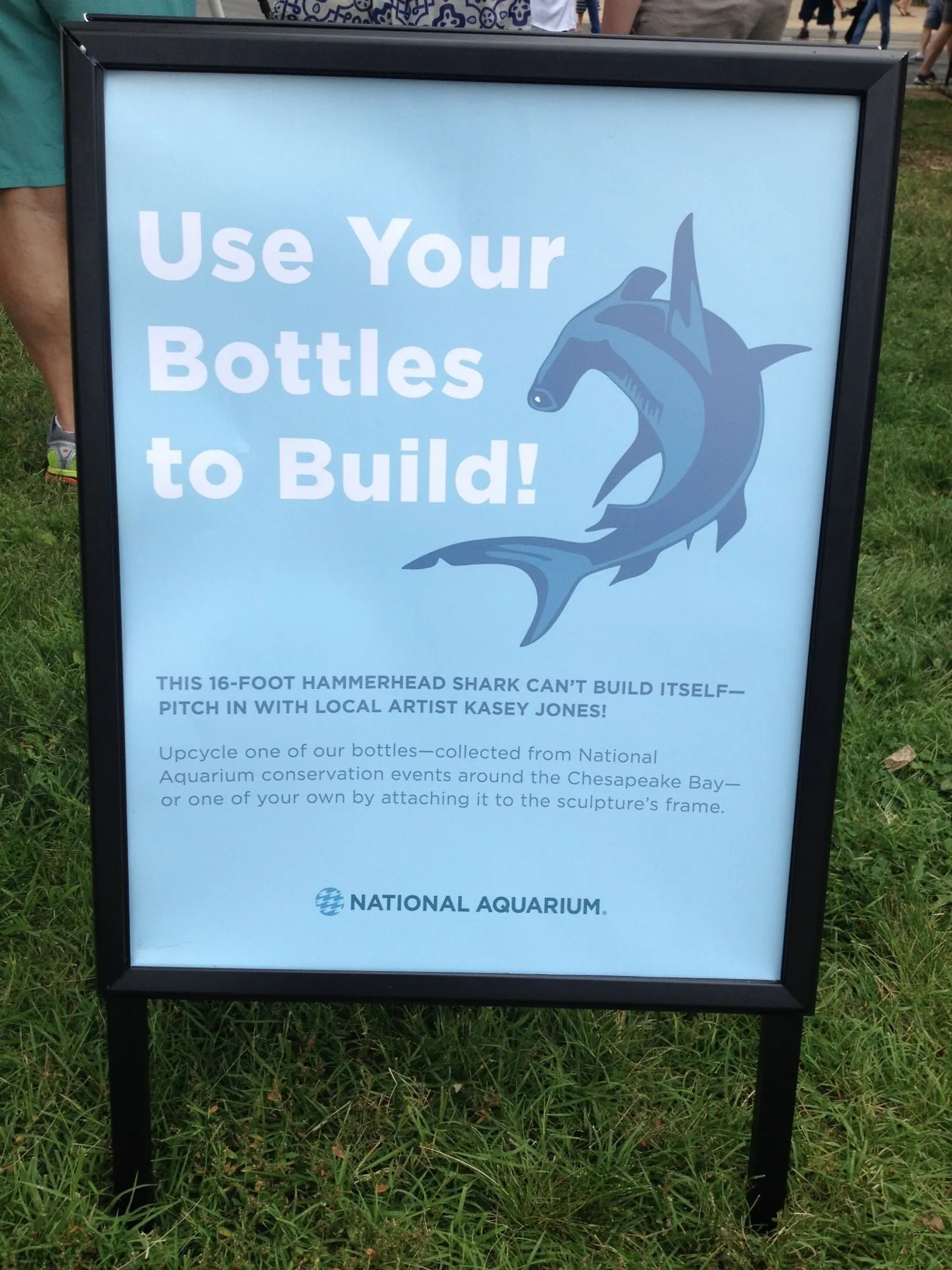 A National Aquarium sign reading "Use Your Bottles to Build! This 16-foot hammerhead shark can't build itself - pitch in with local artist Kasey Jones"