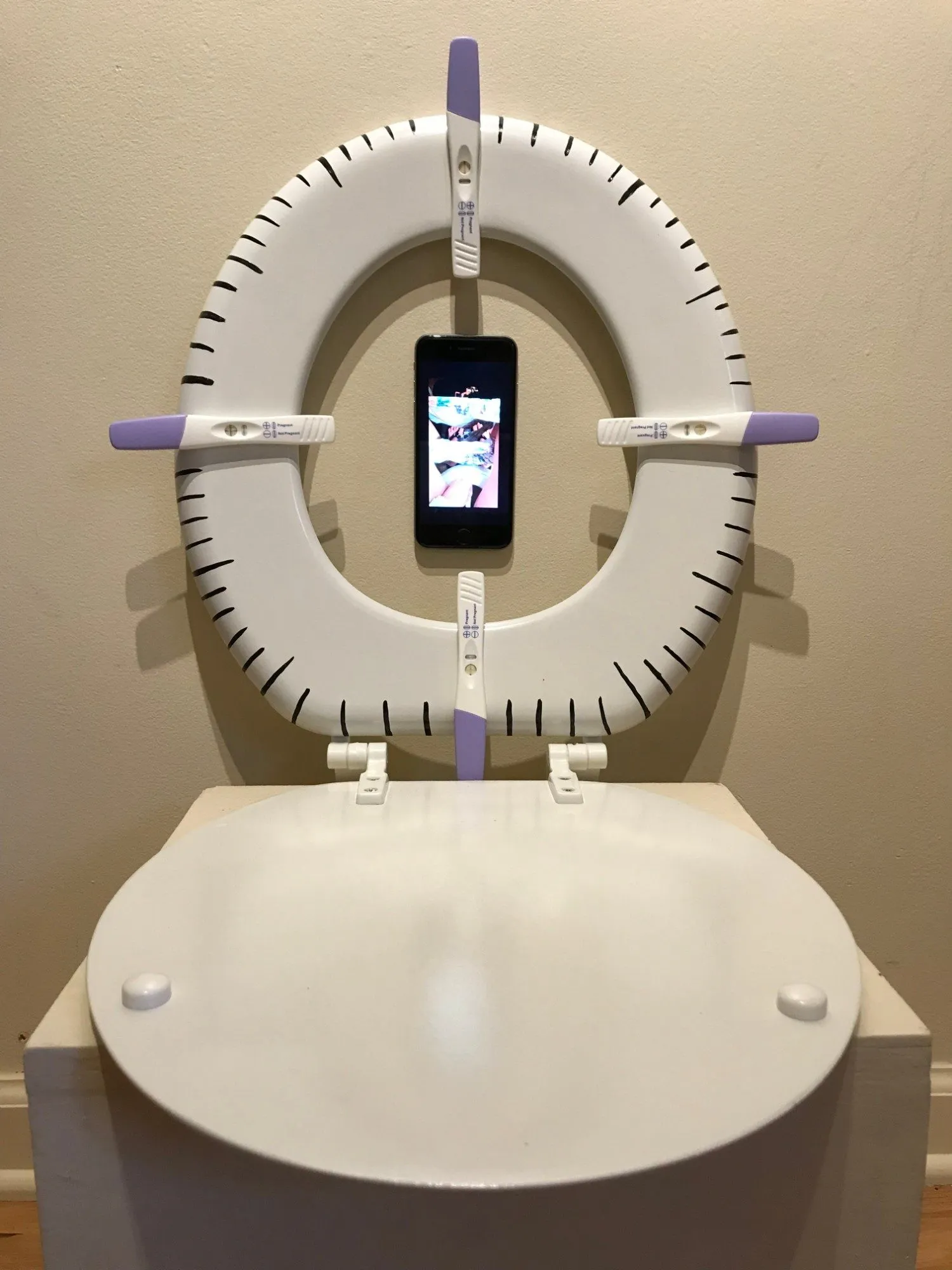 A toilet seat mounted on the wall, four pregnancy tests set into it like the hands of a clock, an iPhone playing video at its center