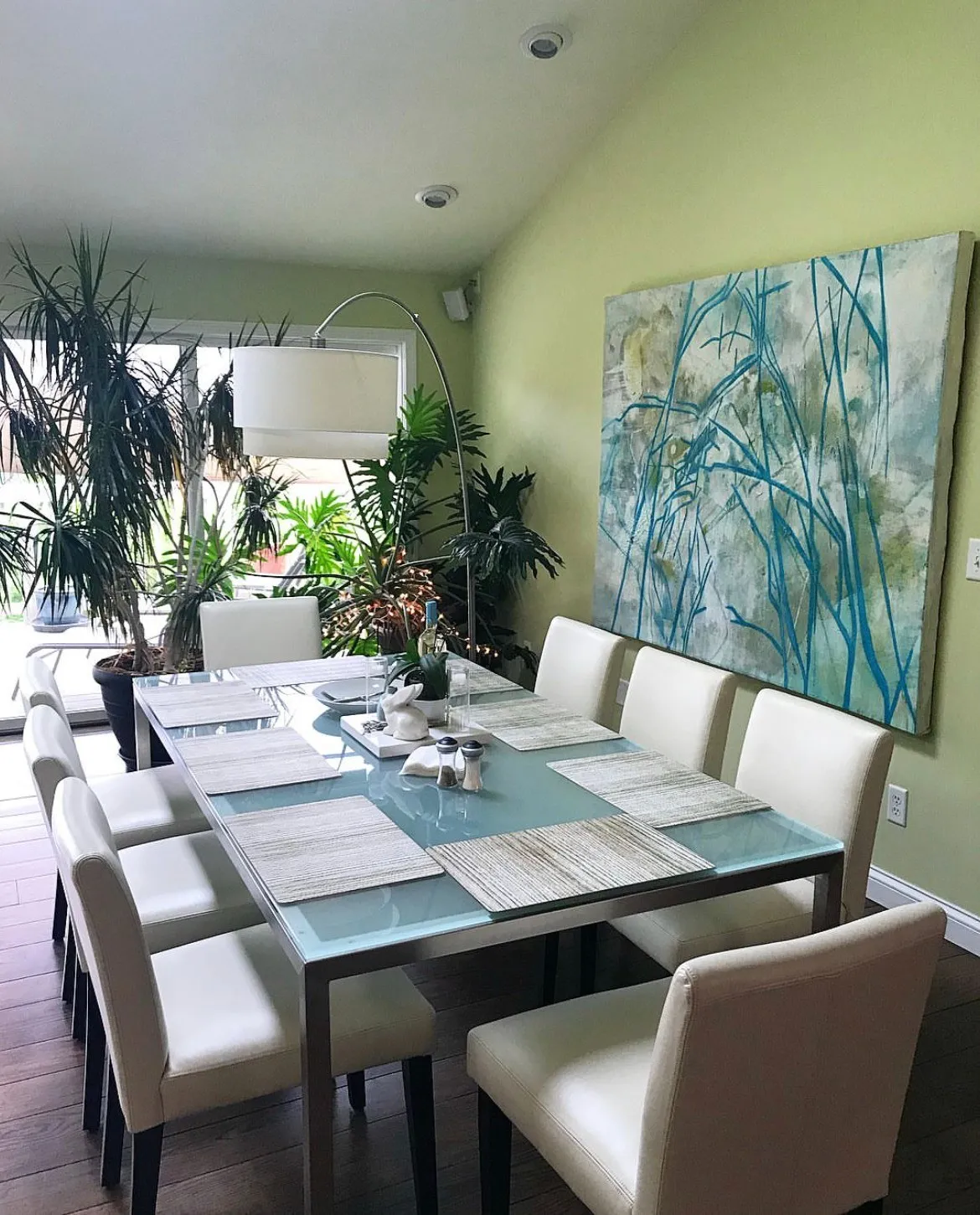 The same painting across the dining room, the long glass table set beneath it and plants massed at the window