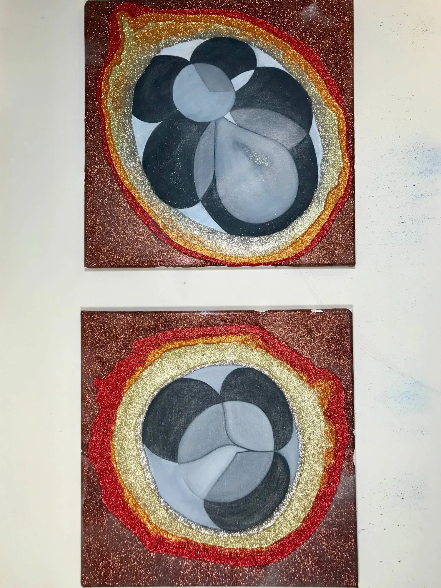 Two square paintings hung one above the other, grey rounded forms ringed by bands of red, gold and silver glitter