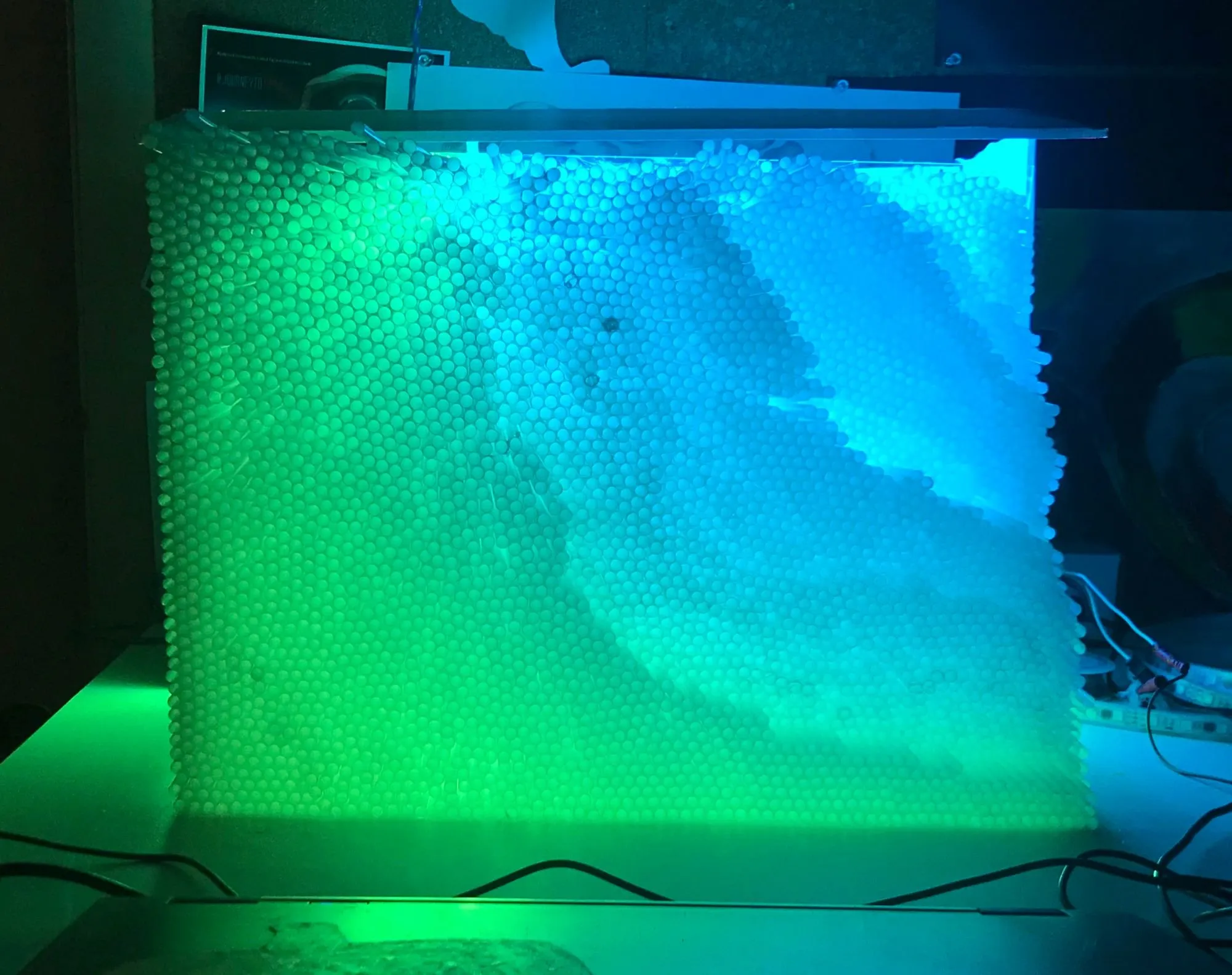 The prototype panel lit from behind, thousands of straw ends glowing green through to blue, LED strips and wiring on the bench beside it