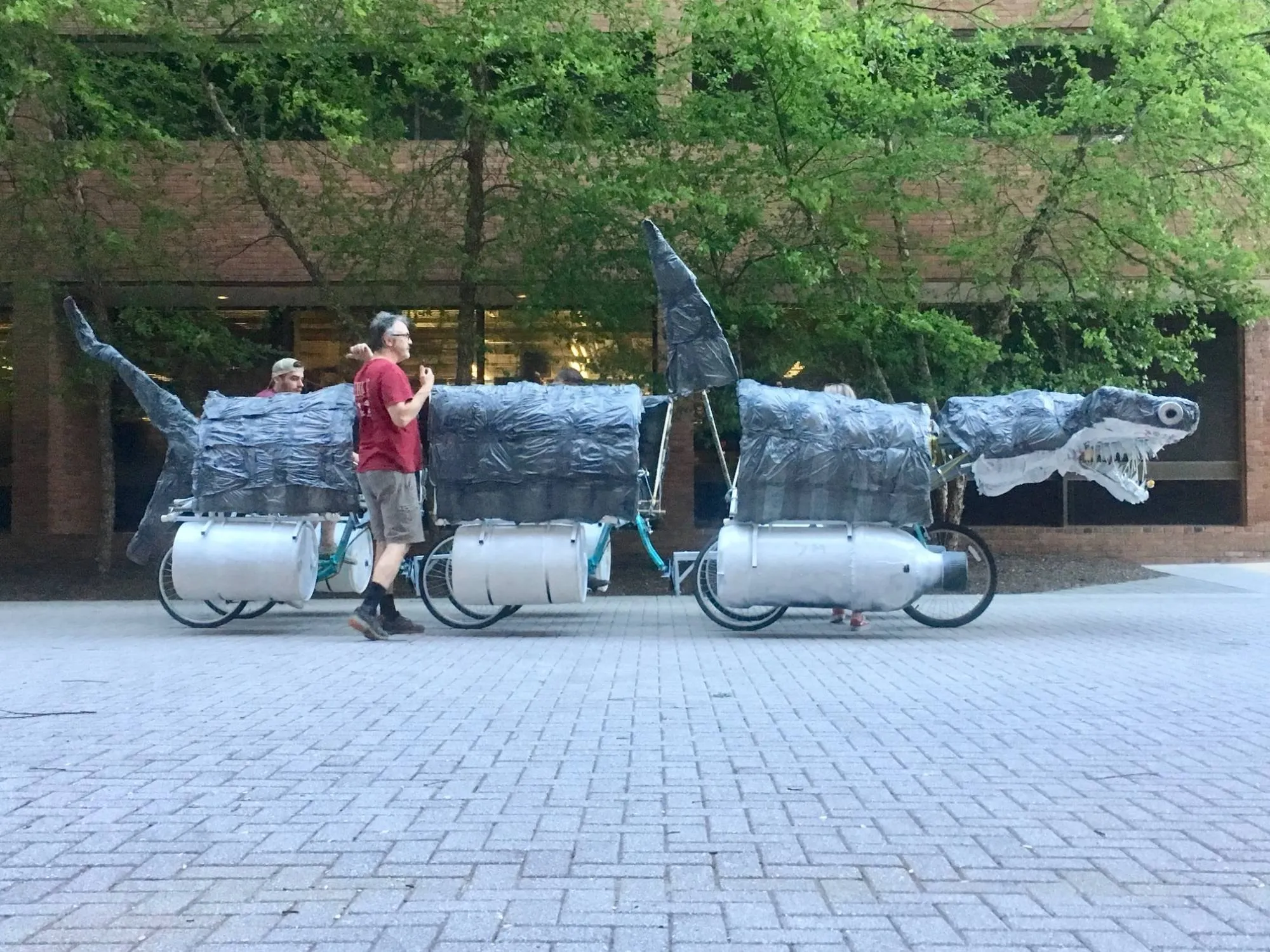 The kinetic shark side on across a brick plaza, its three bodies riding on bottle-shaped floats above the bicycle frames