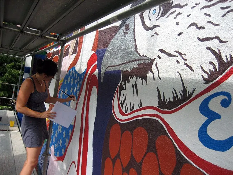 Kasey painting a mural of a large face on an outdoor wall, brush in hand and a reference sheet under her arm