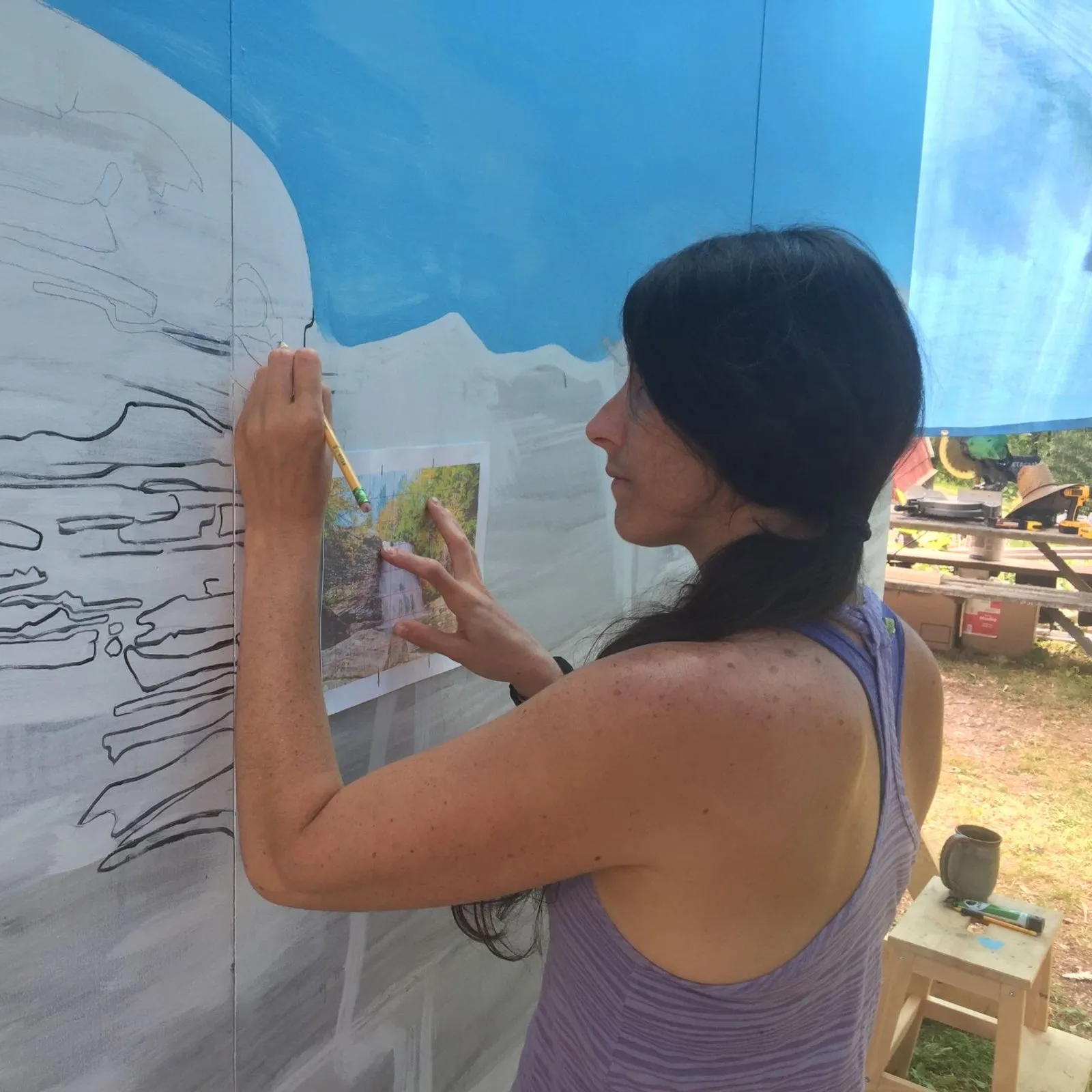Kasey drawing the Swallow Falls mural in pencil, a reference photograph held against the wall