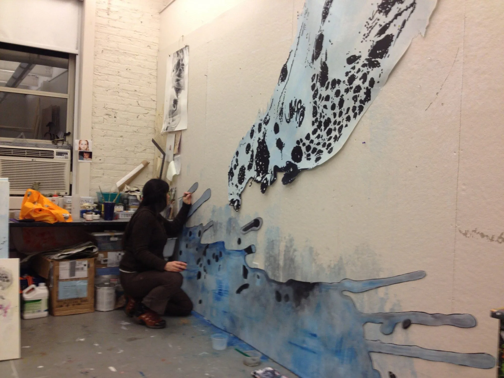 Kasey crouched at the studio wall, painting a large blue and black form that spreads across it